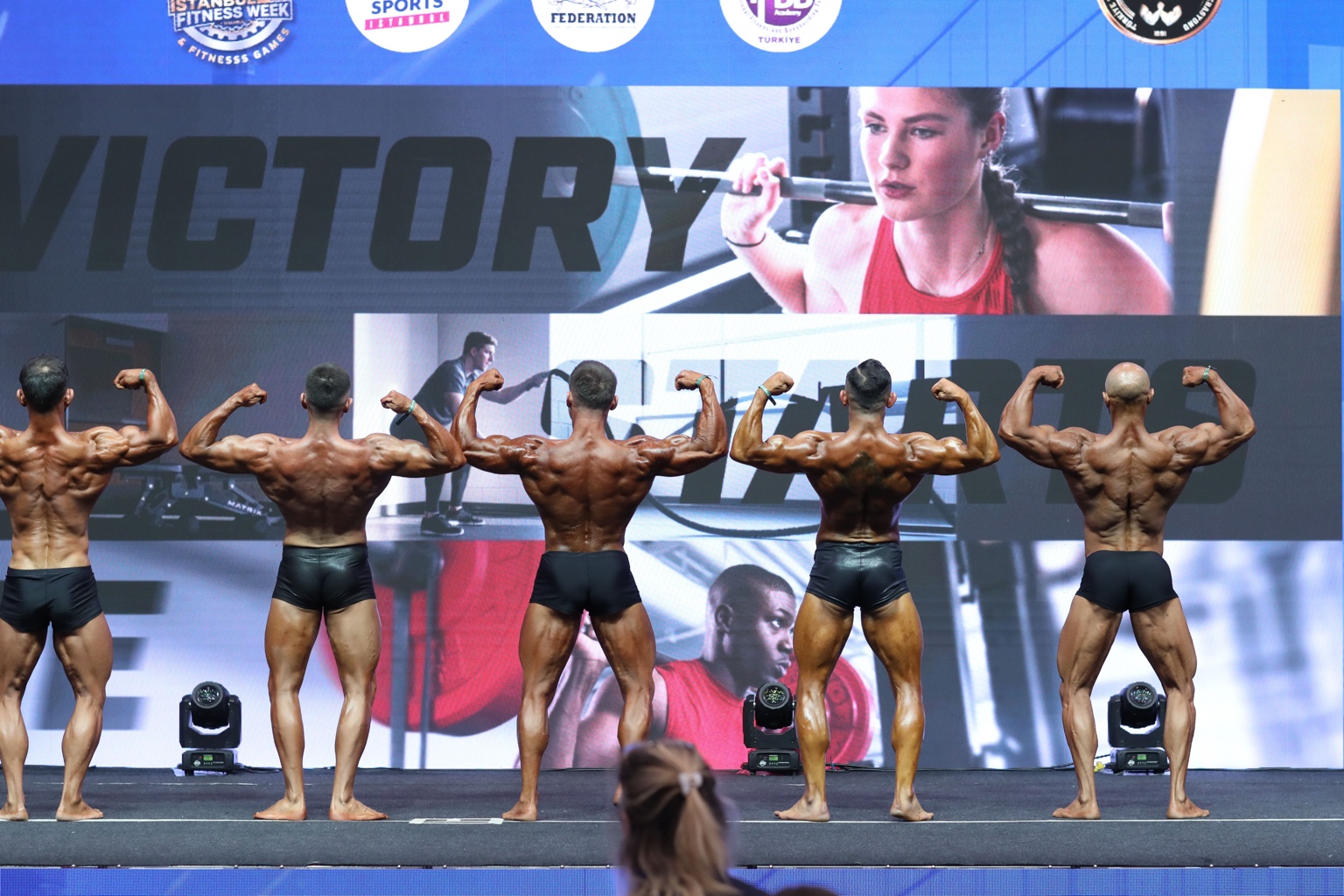 Classic Physique sporcuları sahnede sırt pozu verirken